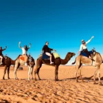 Travelers riding camels across the Sahara sand dunes in Merzouga, Morocco Alt: Camel caravan experience in Merzouga desert with blue sky and golden dunes