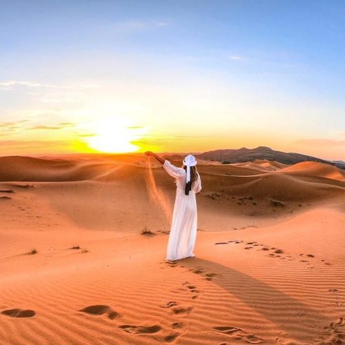 A woman in a white dress stands gracefully in the desert at sunset, capturing the essence of the 5 Days Marrakech Desert Tour.